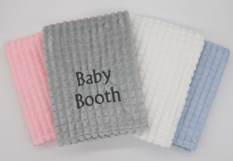 May include: Four baby blankets in different colours: pink, grey, white and blue. The grey blanket has the text "Baby Booth" embroidered on it.