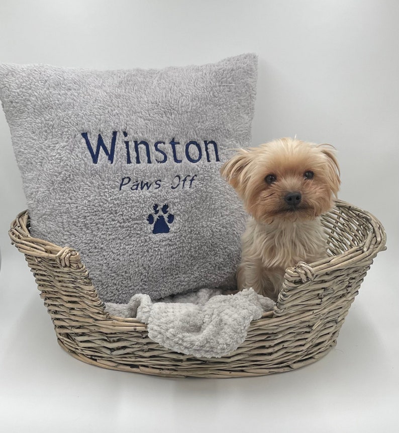 May include: A gray and white dog bed with a gray pillow that says "Winston Paws Off" with a paw print. A small, light brown Yorkshire Terrier dog is sitting in the bed.