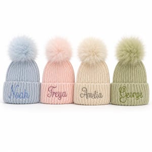 May include: Four ribbed knit beanies with large faux fur pom-poms, each featuring a different embroidered name in cursive script. The hats are light blue with the name Noah, light pink with Freya, cream with Amelia, and sage green with George.