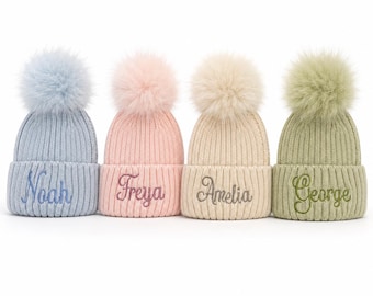 May include: Four ribbed knit beanies with large faux fur pom-poms, each featuring a different embroidered name in cursive script. The hats are light blue with the name Noah, light pink with Freya, cream with Amelia, and sage green with George.