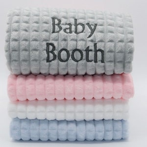 May include: A stack of four plush baby blankets in grey, pink, white, and light blue. The top blanket is grey with the text "Baby Booth" embroidered in grey.
