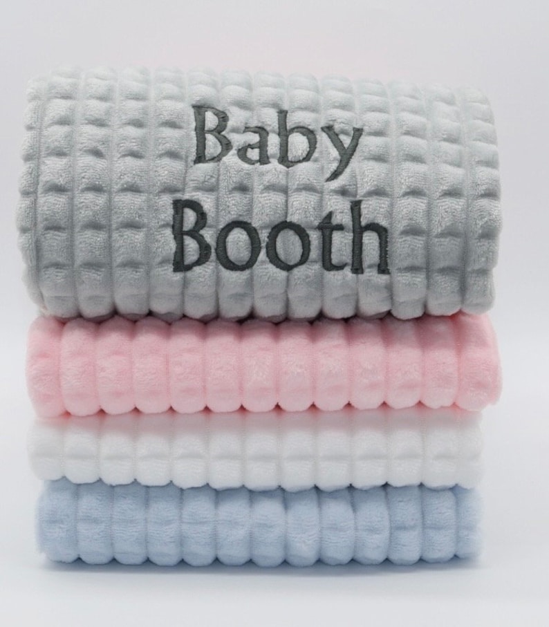 May include: A stack of four plush baby blankets in grey, pink, white, and light blue. The top blanket is grey with the text "Baby Booth" embroidered in grey.