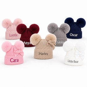 May include: A collection of six ribbed knit beanies, each featuring two large pom-poms on top. The hats come in pink, burgundy, beige, grey, navy, and white. Each beanie is personalised with embroidered names: Lottie, Anya, Harley, Oakley, Oscar, and Little Bear. Some hats include a decorative side bow.