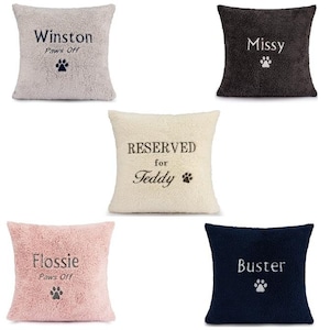 May include: Five different plush dog bed cushions with embroidered names and paw prints. The cushions are in various colors, including beige, pink, brown, white, and blue.