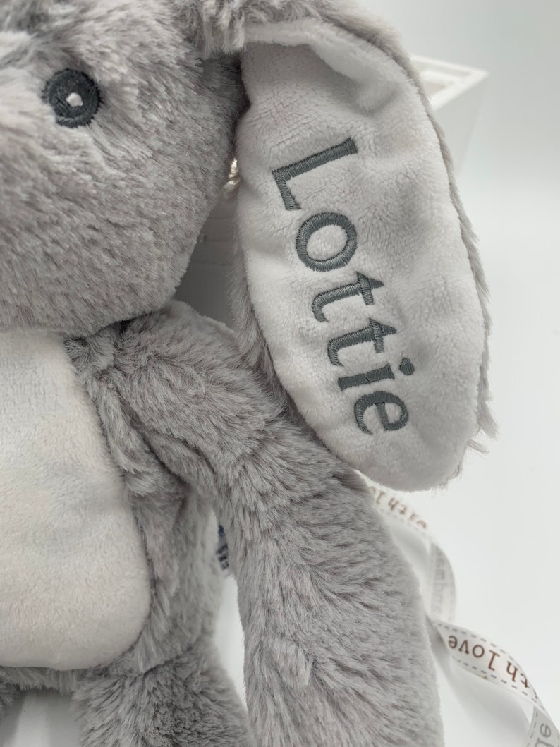May include: A grey plush bunny with a white ear that says "Lottie" embroidered on it. The bunny is holding a white ribbon with the text "with love" on it.