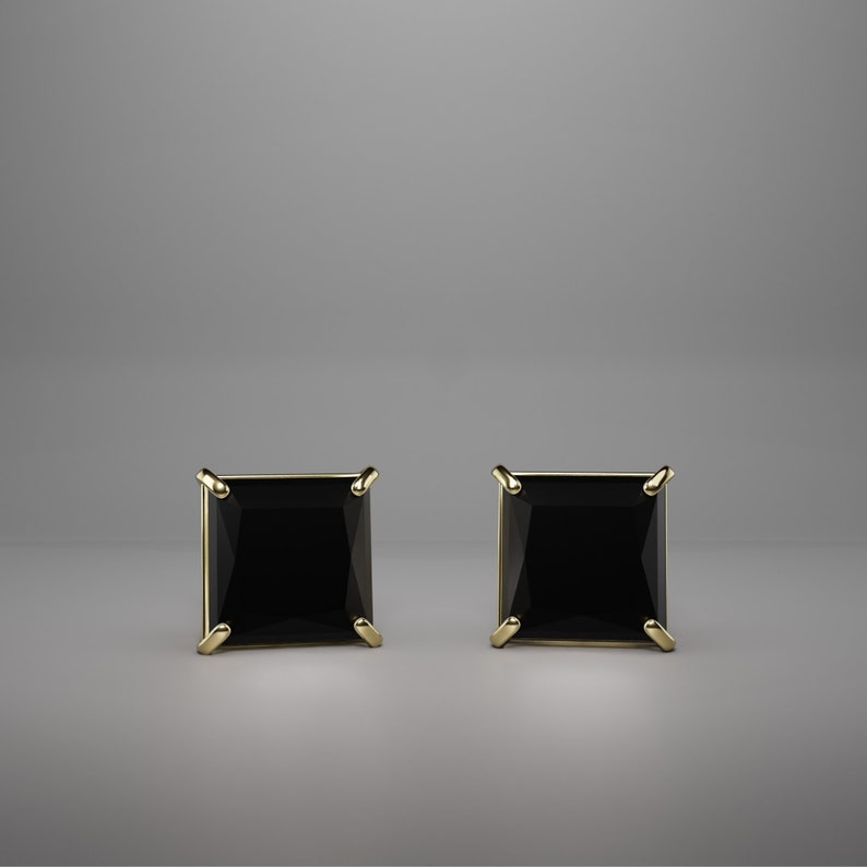Pu&ograve; includere: Un paio di orecchini a lobo con gemme nere a taglio quadrato incastonate in griffe di metallo dorato. Ogni orecchino presenta una pietra scura sfaccettata con una silhouette geometrica netta, fissata da quattro griffe metalliche angolari.