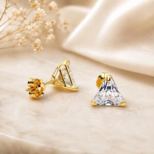 May include: A pair of gold-colored stud earrings with a triangular, clear gemstone. The earrings are set in a gold frame and have a post and butterfly backing. The earrings are displayed on a marble surface.