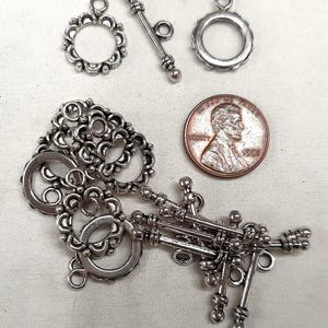 May include: Silver-toned jewelry clasps and components, including toggle clasps and decorative circular pieces. A US penny is included for size reference. The clasps have intricate floral and beaded designs, suitable for jewelry making.