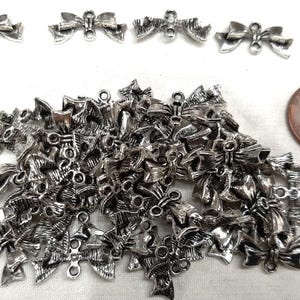 May include: A collection of small, silver-colored bow charms with a loop for attaching to jewelry or crafts. Several bows are scattered, with a penny for size comparison. The bows have a detailed, textured design.