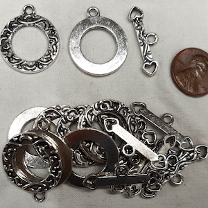 May include: Silver-toned jewelry findings, including ornate round pendants, toggle clasps with heart details, and a penny for size reference. The pendants have a decorative scrollwork design. Ideal for jewelry making.