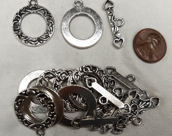 May include: Silver-coloured jewellery findings, including ornate round pendants, toggle clasps with heart details, and a penny for size reference. The pendants have a decorative scrollwork design. Ideal for jewellery making.