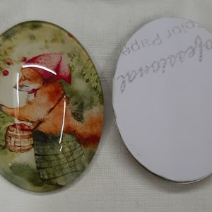Puede incluir: Un cabujón redondo de vidrio con una imagen pintada de un zorro con un sombrero rojo y sosteniendo una cesta de manzanas. El zorro está rodeado de follaje verde y manzanas rojas. La parte posterior del cabujón es blanca con escritura negra.