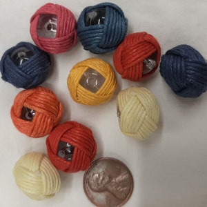 May include: Assortment of ten colorful, woven, round, decorative buttons with a clear plastic center. The buttons are in shades of red, orange, yellow, blue, and cream.