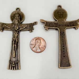 Op de afbeelding: Twee bronzen metalen kruisbeelden. Het kruisbeeld links toont een figuur van Jezus Christus aan het kruis. Het kruisbeeld rechts heeft de woorden "Alpha" en "Omega" op de dwarsbalk. Beide kruisbeelden zijn ongeveer zo groot als een Nederlandse cent.