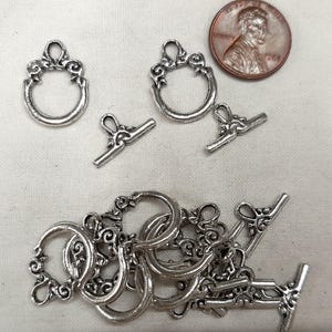 Puede incluir: Cierres de palanca de metal plateado con detalles ornamentados de volutas. Los cierres incluyen anillos circulares y barras en forma de T. Se incluye una moneda como referencia de tamaño.