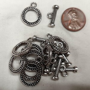 May include: A selection of silver-coloured toggle clasps and rings, with a penny for size comparison. The clasps feature a decorative beaded design around the edges. The rings are also silver and have a similar beaded design.