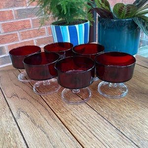 May include: Six vintage ruby red glass dessert cups with clear glass stems. The cups are arranged on a wooden surface, with green plants and a brick wall in the background. The cups are likely from the mid-century.