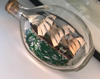 Op de afbeelding: Een glazen fles met een kurkstop die een miniatuur schip met witte zeilen en een groene basis bevat. Het schip is omgeven door een groene en witte vloeistof.