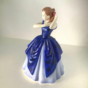 May include: A porcelain figurine of a woman in a blue dress with black detailing, her hair styled up. The dress features a layered design with a light blue underskirt. A decorative item.