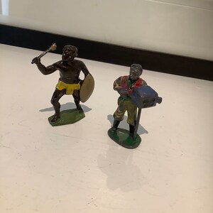 May include: Two painted metal figurines, one holding a spear and shield, the other holding a musical instrument. The figurines are standing on a white surface.