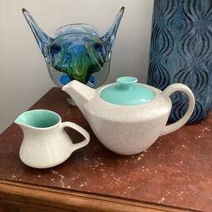 May include: A white teapot with a turquoise lid and a matching creamer are displayed on a wooden surface. A blue and green glass sculpture and a tall blue vase with a textured pattern add to the vintage feel.