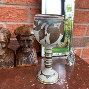 May include: A ceramic goblet featuring a speckled off-white base with hand-painted brown and blue leaf motifs. The stem has a tiered, ringed design leading to a circular foot, with brown painted accents around the base and rim of the cup.