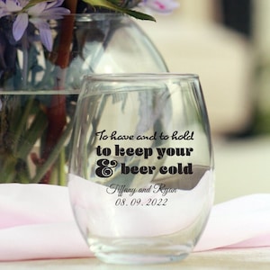 May include: Stemless wine glass with a black and white graphic that says "To have and to hold to keep your & beer cold". The glass is sitting on a white surface with a bouquet of flowers in the background.