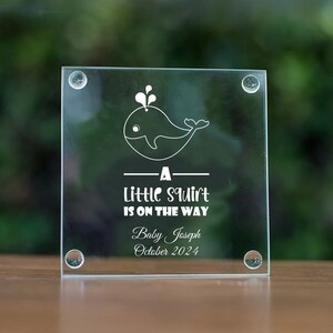 May include: A clear glass coaster with a white whale design and the text "A little squirt is on the way" with the name "Baby Joseph" and the date "October 2024" underneath.