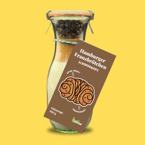 Könnte beinhalten: Ein durchsichtiges Glas mit Metallbügelverschluss enthält geschichtete trockene Zutaten, darunter dunkle Schokodrops oben. Ein braunes rechteckiges Etikett ist befestigt, mit dem Text Hamburger Franzbrötchen Schokodrops, einer Gebäckgrafik und der Angabe Füllmenge 450 g.