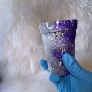 May include: A purple and silver glitter ashtray with a white interior. The ashtray is made of resin and has a round shape.