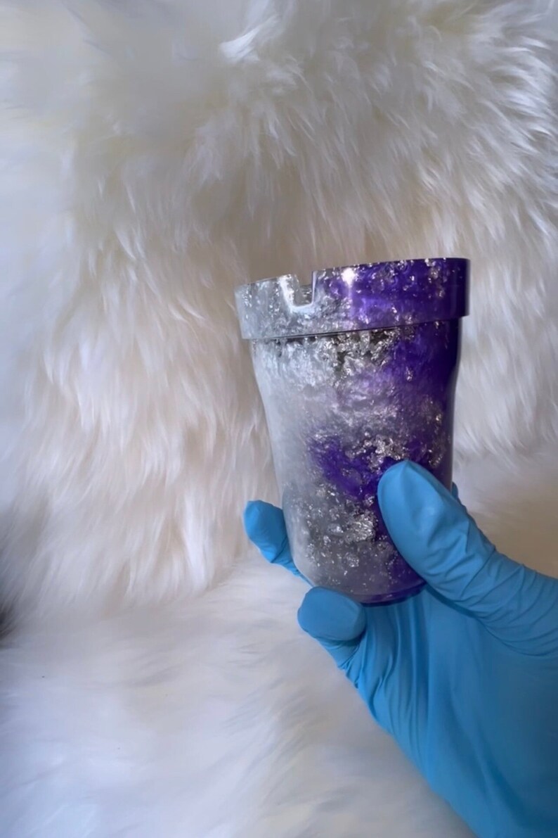 May include: A purple and silver glitter ashtray with a white interior. The ashtray is made of resin and has a round shape.