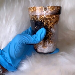 May include: A black and white resin ashtray with gold flecks. The ashtray is shaped like a cup and has a wide rim.