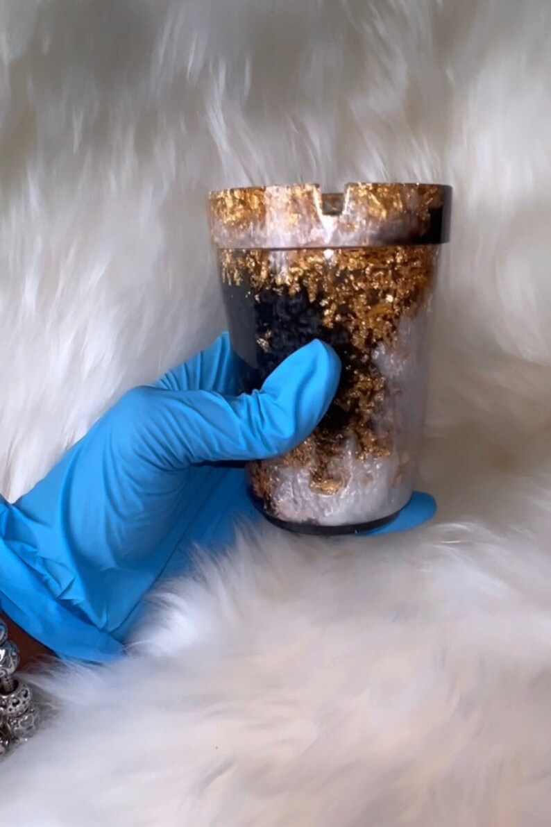May include: A black and white resin ashtray with gold flecks. The ashtray is shaped like a cup and has a wide rim.