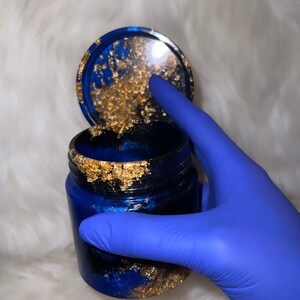 Könnte beinhalten: Ein blaues und goldenes Harzgefäß mit Deckel. Das Gefäß hat ein Wirbeldesign und goldene Flocken durchgehend. Das Gefäß wird von einer behandschuhten Hand gehalten.