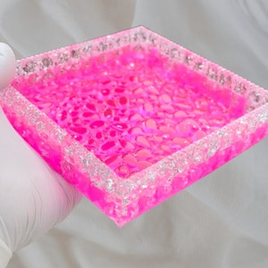 Puede incluir: Una bandeja cuadrada rosa con un borde transparente y texturizado. La bandeja está llena de pequeñas cuentas rosadas translúcidas.