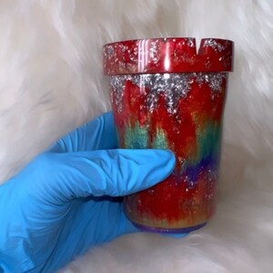 May include: A red, green, and purple tie-dye ashtray with silver glitter accents. The ashtray is made of resin and has a round shape.