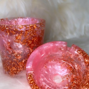May include: A pink and white resin ashtray with gold flecks. The ashtray has a round shape with a small hole in the center.