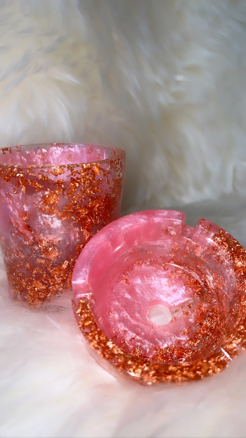May include: A pink and white resin ashtray with gold flecks. The ashtray has a round shape with a small hole in the center.