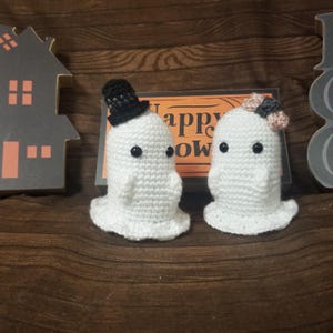 Pode incluir: Duas figuras de fantasmas de crochê brancas com olhos de contas pretas. O fantasma da esquerda usa uma pequena cartola preta e o da direita tem um pequeno laço rosa e cinza. Atrás deles há uma placa retangular com o texto Happy Halloween.