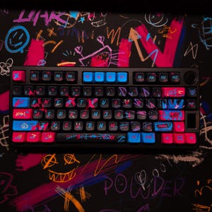 Op de afbeelding: Een zwart mechanisch toetsenbord met aangepaste toetsen in blauw en roze. De toetsen hebben graffiti-achtige ontwerpen en tekst. Het toetsenbord ligt op een zwarte muismat met kleurrijke graffiti-kunst en het woord "POWDER".