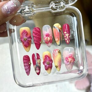 Peut inclure: Un étui en plastique transparent contenant dix faux ongles. Les ongles sont décorés de fleurs et de volutes roses, blanches et jaunes.