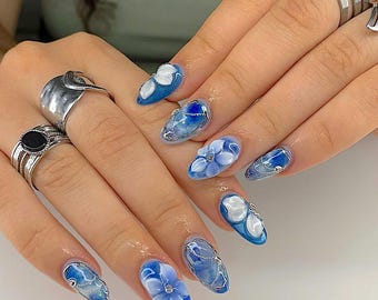 May include: Oval-shaped fingernails with a blue and white floral design. The nails feature 3D flower accents, silver chain details, and a glossy finish. The design incorporates shades of blue and white, creating a delicate and artistic look.