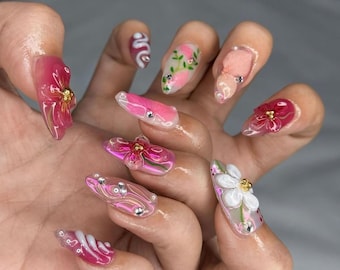 Puede incluir: Una mano que muestra una manicura con uñas acrílicas largas y transparentes con varios diseños. Las uñas presentan adornos florales 3D en rosa y blanco, pedrería y patrones abstractos. Algunas uñas tienen un diseño de serpiente.