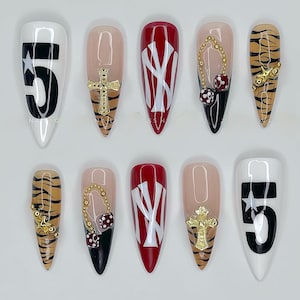 May include: A collection of ten long, pointed artificial nails. Designs include a white nail with a black number 5 and star, a red nail with a white logo, and nails with gold cross, dice, and tiger stripe patterns.