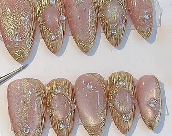 May include: A set of ten artificial nails in various shapes. The nails feature a combination of gold and pink hues, with some having a textured gold design and others a smooth pink finish. Each nail is adorned with small, sparkling rhinestones.