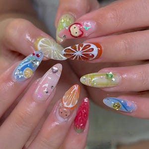May include: A set of long, almond-shaped fingernails with colorful fruit-themed nail art. Designs include a lemon slice, orange slice, strawberries, and cartoon characters. Some nails feature glitter and star-shaped accents.