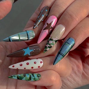 May include: A hand displays a set of long, pointed acrylic nails with various designs. The nails feature patterns like polka dots, stars, camouflage, and plaid in colors such as brown, pink, blue, green, and white. The nails are well-manicured and showcase a variety of nail art.