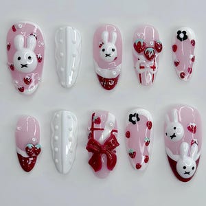 May include: A set of ten artificial nails with a pink base, featuring white bunny and strawberry designs. Some nails have 3D elements like bows and strawberries. The nails are almond-shaped.