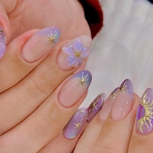 May include: A set of manicured nails with a gradient design in shades of purple and pink. Each nail features gold star accents and a 3D flower. The nails are oval-shaped.