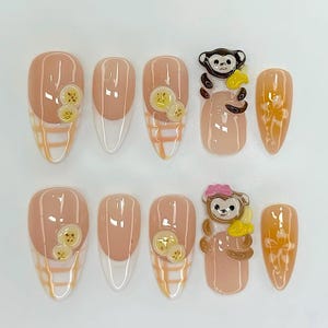 May include: A collection of ten artificial nails in a light peach hue. Some nails have a white tip, others a yellow and white checked pattern. Decorations include banana slices and monkey figures, creating a fun, whimsical design. (Measurements are not applicable).